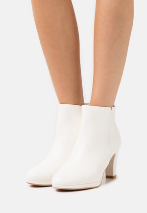 Chevilles portant des bottines blanches en cuir à talons hauts avec bouts arrondis sur un fond uni.