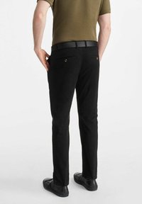 Osoba stoi tyłem, ubrana w dopasowaną oliwkową koszulę, czarne spodnie i czarne buty, z rękami opartymi na biodrach.