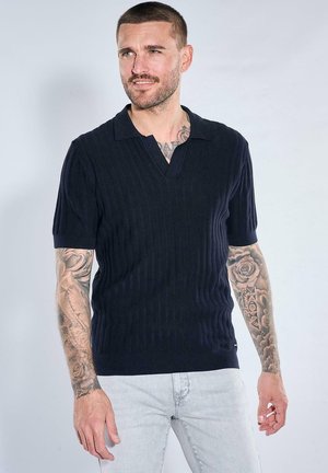 Man with tattoos wearing a black short-sleeve ribbed polo shirt and light gray jeans, standing against a plain background.