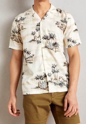 Homme portant une chemise beige à manches courtes avec une impression de palmiers tropicaux et de planches de surf, associée à un short marron.