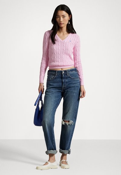 Pink cable-knit sweater with V-neck, paired with distressed blue jeans. Model holds a blue handbag and wears beige slip-on shoes.