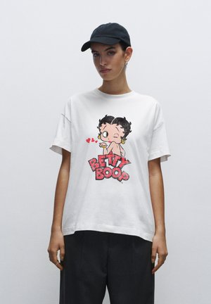 Hvid bomulds t-shirt med et farverigt Betty Boop grafik med rød tekst, rund halsudskæring og korte ærmer. Kombineret med mørke bukser.