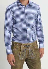 Mann in einem blau-weißen karierten Hemd mit Knöpfen, das in eine traditionelle braune Lederhose mit gestickten Details gesteckt ist.
