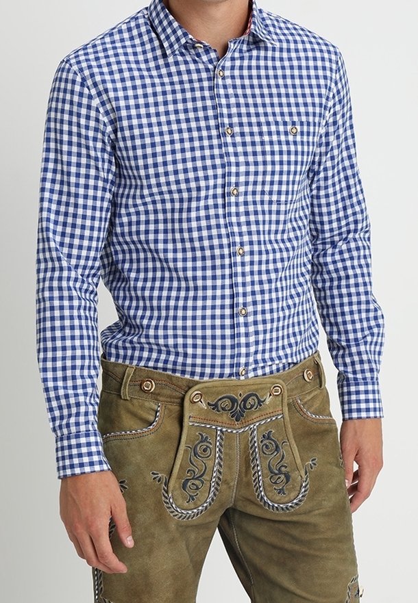 Mann in einem blau-weißen karierten Hemd mit Knöpfen, das in eine traditionelle braune Lederhose mit gestickten Details gesteckt ist.