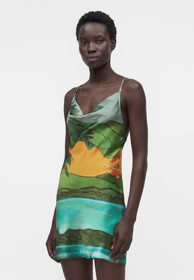 Γυναίκα που φοράει ένα αμάνικο φόρεμα με λεπτές τιράντες με πολύχρωμο τροπικό τοπίο που περιλαμβάνει νερό, βλάστηση, πορτοκαλί ουρανό και πουλιά.