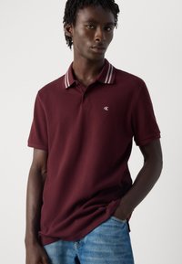 Jonge man met korte dreadlocks, gekleed in een bordeauxrode polo met gestreepte kraag en blauwe spijkerbroek, die poseert met één hand in zijn zak.