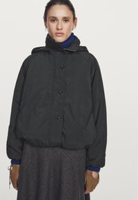 Zwarte gewatteerde jas met een capuchon, voorzien van knoopsluitingen, gedragen over een bruin geruit rokje. Handschoenen van donkerbruin leer met uitgesneden vingers.