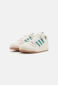 Låga vita sneakers med teal Adidas-ränder, beige sulor, guld Adidas-logotyp och snörning på en vit bakgrund.