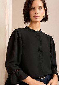 Black blouse with a scalloped neckline, pleated front, and puff sleeves with lace trim. Soft fabric and flared cuffs enhance the design.