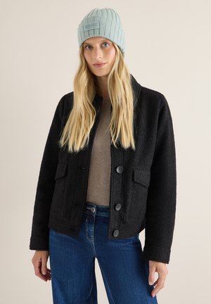 Black textured jacket with button closure and two front pockets, paired with a light blue ribbed beanie and beige top.