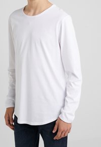 T-shirt à manches longues blanc en coton doux, avec un col ras du cou et un petit logo en relief sur la poitrine, associé à un jean bleu.