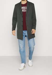 Grau melierter Wollmantel mit strukturiertem Design, ausgestattet mit einer vorderen Knopfverschluss, getragen über einem burgunderfarbigen Grafik-T-Shirt und hellblauen Jeans.