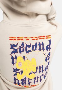 Beige hoodie met een kleurrijke grafische print op de achterkant. Het ontwerp bevat een speelse gele cartoonhond en gedurfde, gestileerde tekstelementen.