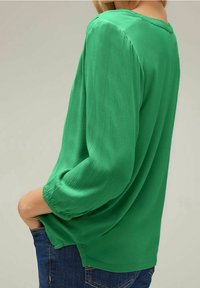 Blouse verte, légère, avec de longues manches légèrement bouffantes. Texture lisse, encolure arrondie et fentes latérales pour faciliter le mouvement.