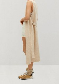 Długi dzianinowy bezrękawnik w miękkim odcieniu beżu z wiązaniem z tyłu, nałożony na jasną zieloną bluzkę i białe szorty, zestawiony z beżowo-żółtymi sneakersami.