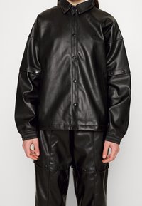 Svart imitationläderjacka med knappstängning framtill, spetsig krage och elastiska ärmslut. Stylar med matchande byxor, visar en slank textur.