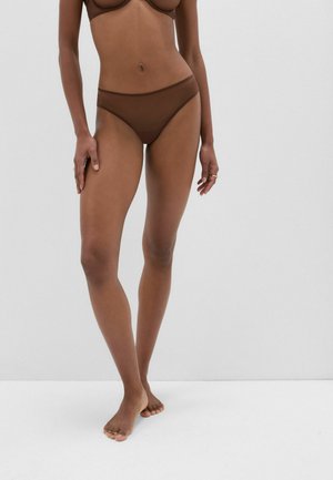 Vrouw staat blootsvoets, draagt een bruine beha en bijpassend laag uitgesneden ondergoed tegen een effen witte achtergrond.