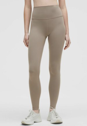 Leggings beige a vita alta realizzati in tessuto liscio e elastico, con un design aderente e cuciture senza soluzione di continuità. Indossati con sneakers chiare.