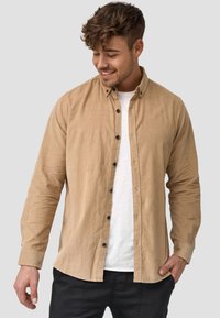 Camisa de botón marrón claro con una tela texturizada, dos bolsillos frontales y botones oscuros. Usada sobre una camiseta blanca y combinada con pantalones negros.