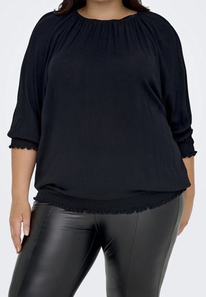 Kobieta w luźnej czarnej bluzce z marszczonym dekoltem i mankietami, zestawionej z dopasowanymi czarnymi skórzanymi spodniami na jednolitym tle.