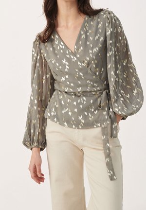 Kobieta ubrana w szarą bluzkę typu wrap z metalicznymi białymi i złotymi plamkami, bufkowymi długimi rękawami oraz wiązaniem z boku, w zestawieniu z kremowymi spodniami.