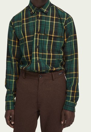 Mann trägt ein grünes, gelbes und schwarzes kariertes Hemd mit Knöpfen, das in hoch taillierte braune Hosen mit Gürtelschlaufen gesteckt ist.