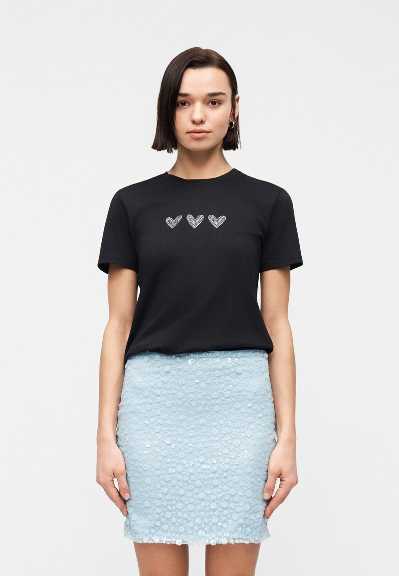 Γυναίκα με κοντά σκούρα μαλλιά που φοράει μαύρο μπλουζάκι με τρεις καρδιές από στρας και ελαφρύ μπλε υφασμάτινο μίνι φούστα, στέκεται μπροστά σε λευκό φόντο.