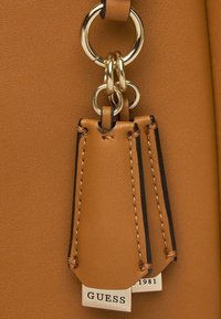 Détail d'un sac à main en cuir tan avec un anneau en or, une étiquette en cuir avec des coutures contrastées et une étiquette de marque.