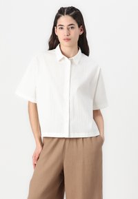 Camisa blanca de manga corta con una tela texturizada, con botones en la parte delantera y un corte relajado, combinada con pantalones marrón claro.