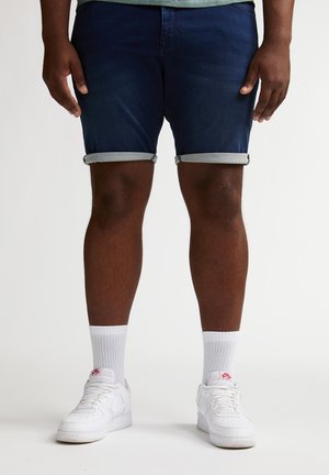 Donkerblauwe denim shorts met opgerolde zoom, gecombineerd met witte sneakers en witte enkelsokken. De shorts hebben strakke lijnen en een gladde textuur.