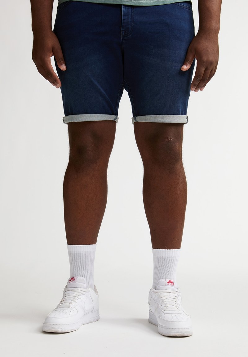 Pantalones cortos de mezclilla azul oscuro con dobladillos doblados, combinados con zapatillas blancas y calcetines blancos hasta el tobillo. Los pantalones cortos presentan líneas limpias y una textura suave.