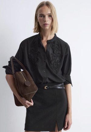 Frau mit kurzem blonden Haar, die eine schwarze bestickte Bluse und einen schwarzen Rock mit Schlitz trägt und eine braune Wildlederhandtasche vor einem weißen Hintergrund hält.