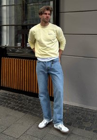 Geel sweatshirt met logo, wit T-shirt eronder, blauwe jeans en witte sneakers met zwarte strepen. Casual urban outfit.