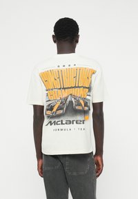 Valge puuvillane T-särk, millel on suur graafiline trükk võidusõiduaudadest ja tekst "2024 CONSTRUCTORS' CHAMPIONS" ja "McLaren Formula 1 Team" oranžis värvuses.