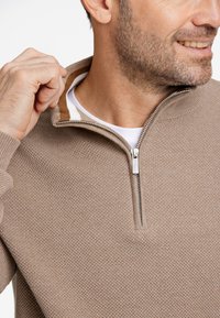 Suéter beige con cremallera y un patrón de punto texturizado, que presenta un cuello a rayas en marrón y blanco en contraste y un tirador de cremallera plateado.