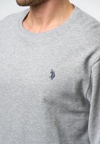 Grå hettegenser laget av mykt stoff, med rund hals og en liten mørkeblå brodert logo på brystet. Enkel, klassisk design.