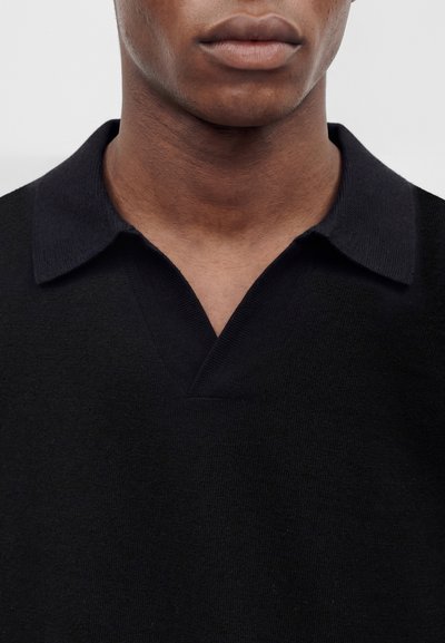 Lèvres et partie inférieure du visage d'une personne portant un pull en maille noir avec un col et un décolleté en V sur un fond uni.