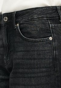 Κοντινή λήψη της μέσης μαύρου τζιν παντελονιού με μπροστινή τσέπη και κουμπί, φορεμένο με άσπρη ριγέ μπλούζα μέσα στο παντελόνι.