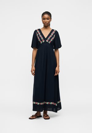 Donna che indossa un abito nero lungo con scollo a V e ricamo floreale rosa sullo scollo e sull'orlo, abbinato a sandali neri.