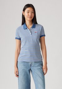 Πολυάρι με μπλε και λευκές ρίγες, με κουμπωτό γιακά, διαθέτει κοντά μανίκια και μικρό κόκκινο λογότυπο στο στήθος. Φοριέται με ανοιχτόχρωμα μπλε τζιν.