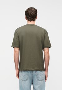 Άνδρας που φοράει κοντομάνικη μπλούζα χακί και ανοιχτόχρωμα τζιν, στεκόμενος με την πλάτη στραμμένη μπροστά σε λευκό απλό φόντο.