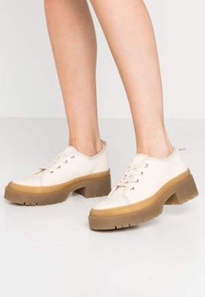 Zapatillas de lona beige con una suela de goma gruesa y translúcida y puntera redondeada. Presentan ojales plateados, cordones y una tira de agarre en el talón.