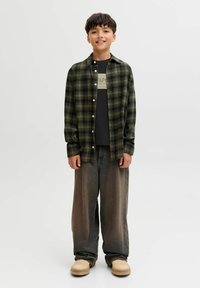 Geruit flanellen overhemd in donkergroen en zwart, gedragen over een zwart T-shirt met witte letters. Loszittende grijze broek en beige schoenen.