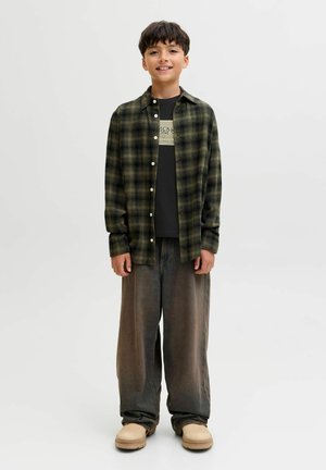 Geruit flanellen overhemd in donkergroen en zwart, gedragen over een zwart T-shirt met witte letters. Loszittende grijze broek en beige schoenen.