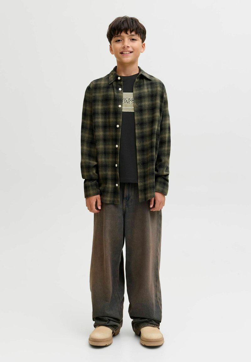 Geruit flanellen overhemd in donkergroen en zwart, gedragen over een zwart T-shirt met witte letters. Loszittende grijze broek en beige schoenen.