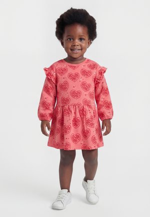 Usmievajúca sa dievčatko, oblečené v ružových šatách s červenými srdiečkami "Howdy Honey" a bielymi teniskami, stojí na jednoduchom pozadí.