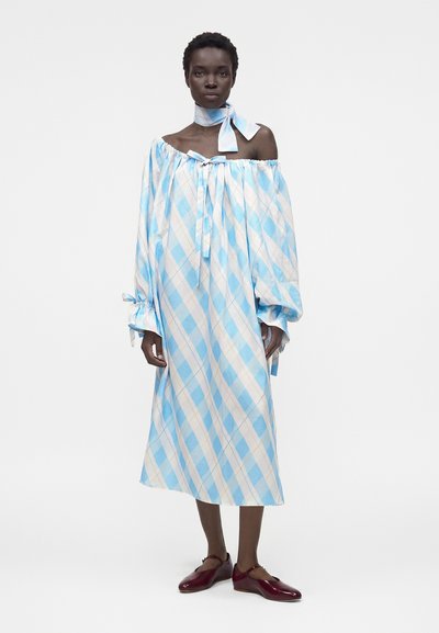 Γυναίκα με αέρινο, καρό φόρεμα σε μπλε και άσπρο χρώμα με μακριά μανίκια και λαιμοκάπα με σχέδιο που ταιριάζει, συνδυασμένο με σκούρα κόκκινα επίπεδα παπούτσια.