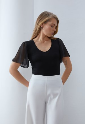 Femme aux longs cheveux blonds portant un haut noir à manches courtes transparentes et un pantalon blanc taille haute, debout les mains derrière le dos.
