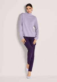 Eine Frau trägt einen helllila Rollkragenpullover, dunkelviolette Hosen und helllila Absätze und steht vor einem schlichten hellen Hintergrund.