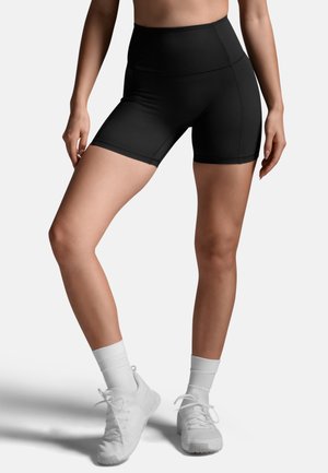 Donna che indossa pantaloncini sportivi aderenti neri a vita alta, calzini bianchi alla caviglia e scarpe da ginnastica bianche, in piedi con le gambe incrociate.